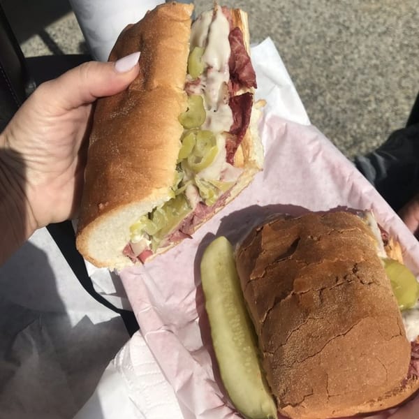 A hand holds a large deli sub sandwich with meat, cheese, and peppers, next to another sub and pickle.